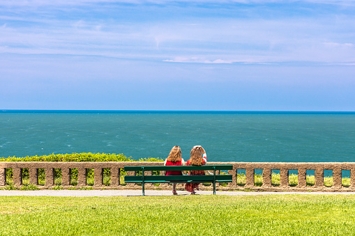 Two twin red head women in red dresses sitting on an isolated bench in front of the Atlantic Ocean in Biarritz, France