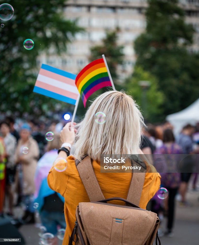 Woman Waving  Rainbow Pride Flags At The Love Festival Rear view of a young blond woman in a yellow shirt walking at the LGBTQIA pride event and waving rainbow flags LGBTQIA Pride Event Stock Photo