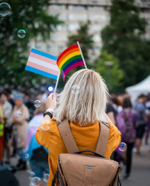 Rear view of a young blond woman in a yellow shirt walking at the LGBTQIA pride event and waving rainbow flags