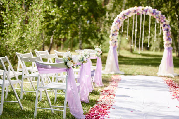 Place for wedding ceremony. Wedding arch decorated with cloth and flowers and chairs on each side of archway outdoors, copy space. Empty wooden chairs for guests on green grass. Wedding setup stock photo