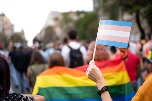 A female hand holding the transgender flag at pride parade with blurred participants in the background
