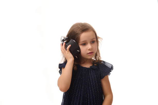 a beautiful curious little girl in a dark blue dress holds an alarm clock near her ear and listens attentively to the sound of the arrows, isolated on white background with copy space - clock clock face blank isolated imagens e fotografias de stock
