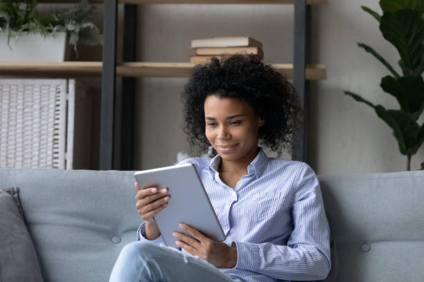 istock Smiling african woman sit on couch enjoy using electronic tablet 1341643936