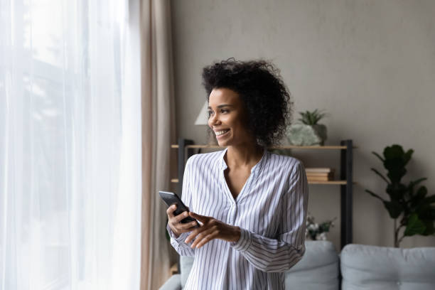 istock Cheerful young black woman using phone near window 1341643997