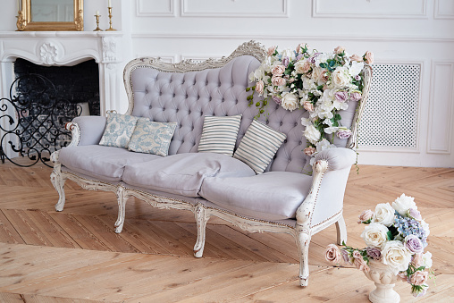Soft sofa with gray fabric upholstery, pillows and flowers, copy space. Luxury rich living room interior with elegant vintage textile couch and fireplace. Chesterfield upholstery