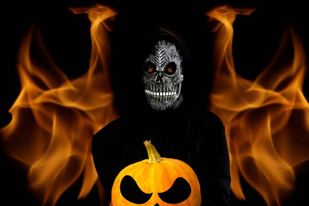 Grim reaper holding crop halloween pumpkin head. Man in death mask with fire flame in eyes and grin on dark black hell fire flame background.Halloween holiday concept.Dark horror. Selective focus Grim reaper holding crop halloween pumpkin head. Man in death mask with fire flame in eyes and grin on dark black hell fire flame background.Halloween holiday concept.Dark horror. Selective focus. halloween pumpkin human face candlelight stock pictures, royalty-free photos & images