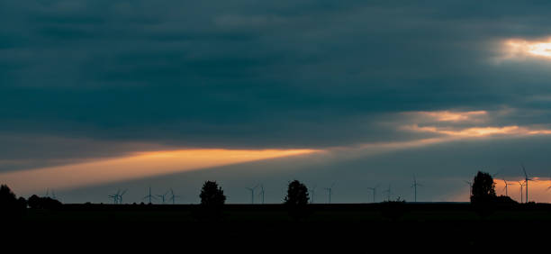 silhouette d’éoliennes et d’arbres à l’horizon au coucher du soleil. ciel spectaculaire et lumineux avec un grand rayon de soleil orange brillant à travers les nuages sombres. - dramatic sky dusk night sustainable resources photos et images de collection