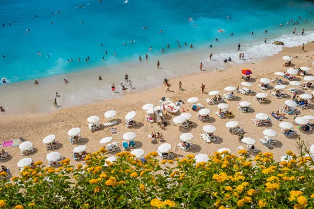 The sea with a variety of turquoise and dark blue colors. The beach on a steep extreme land. There are many tourists.In the first layer there are flowers beach and sea.Tourists enjoying the beach with lots of sunbeds and umbrellas