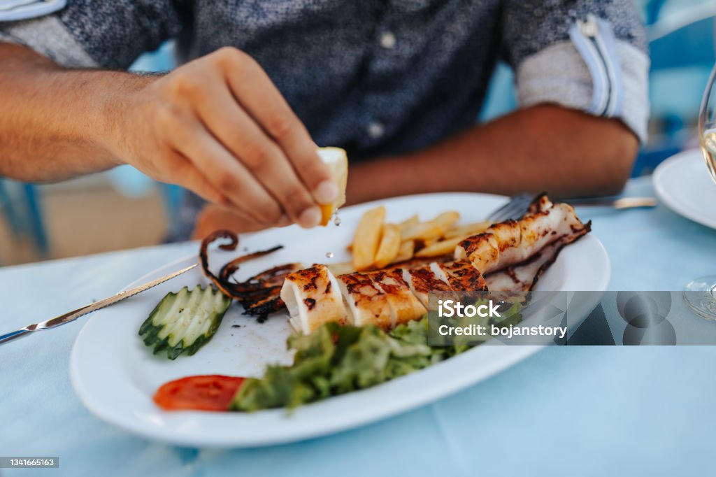 The secret is in the spices Close-up shot of young male hand squeezes a lemon on a grilled squid at outdoor restaurant Fish Stock Photo
