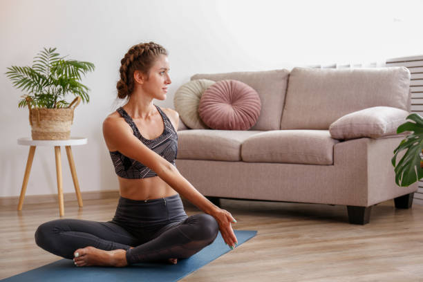 Yogini exercising at home Sporty young woman practicing yoga in the comfort of her own home. Yogini performing morning physical exercise routine at the living room. Interior background, copy space, close up. sukhasana stock pictures, royalty-free photos & images