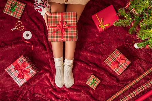 Top view of woman with christmas gift box on knees. Female wrapping gifts for family sitting on fluffy plaid near christmas tree. Flat lay.