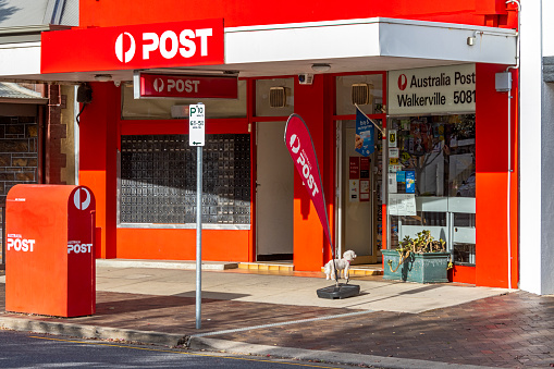 Adelaide, Australia - May 5, 2021: Walkerville 5081 Post Office, Walkerville Tce; a small white dog on a lead waits at the open front door while its owner collects the mail from inside the post office. Very bright red facade, three-quarter front view showing Australia Post signage, secure private mail boxes, posting box and advertising banner, open front door.