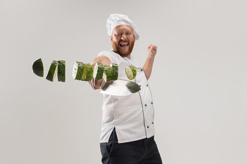 Unique cutting of vegetables. One hadsome bearded man, cook, male chef in white uniform cut cucumber isolated on white background. Cuisine, profession, occupation, funny meme emotions concept.