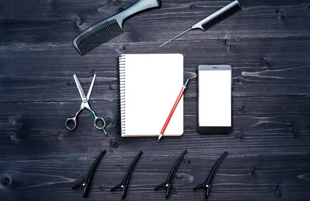 Hairdresser tools on wooden background. Blank card with barber tools flat lay. Top view on wooden table with scissors, comb and hairclips with empty notebook, pencil and phone, free space