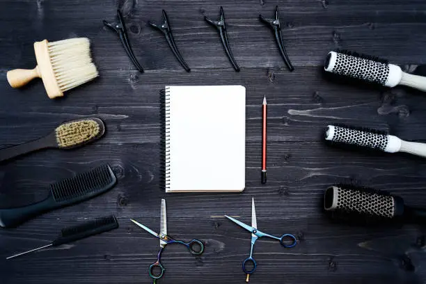 Hairdresser tools on wooden background. Blank card with barber tools flat lay. Top view on wooden table with scissors, comb, hairbrushes and hairclips with empty notebook and pencil, copy space