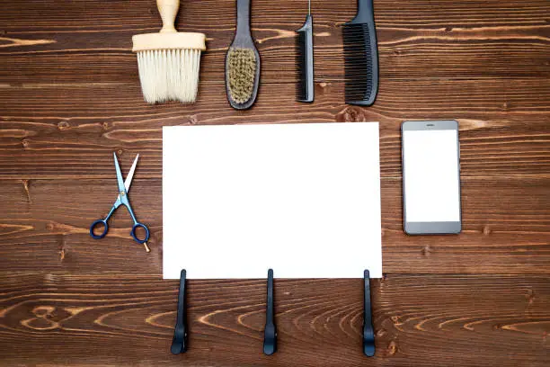 Hairdresser tools on wooden background. Blank card with barber tools flat lay. Top view on wooden table with scissors, comb, brush and hairclips with empty white paper and phone, free space