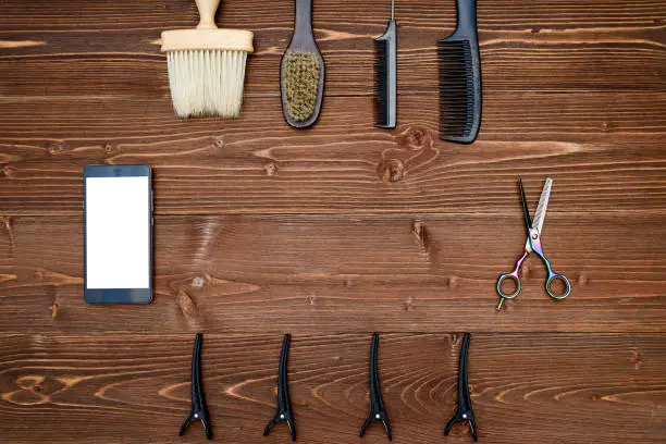 Hairdresser tools on wooden background. Top view on wooden table with scissors, comb, brush and hairclips with phone, free space
