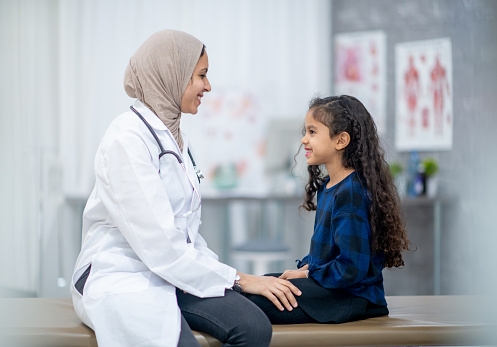 A young girl sits in casual clothing with her legs crisscrossed, on an examination table. She is smiling up at her doctor as they sit close together, face-to-face. Her female Muslim doctor is sitting up on the table in front of her, wearing a while lab coat and a hijab as she rests her hand on the girls knee to reassure her.