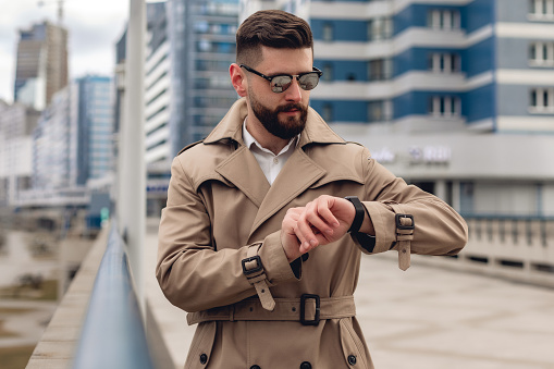 Portrait of bearded caucasian man in beige jacket and white shirt in the city. Business concept. Caucasian guy with fashion black sunglasses who follow time
