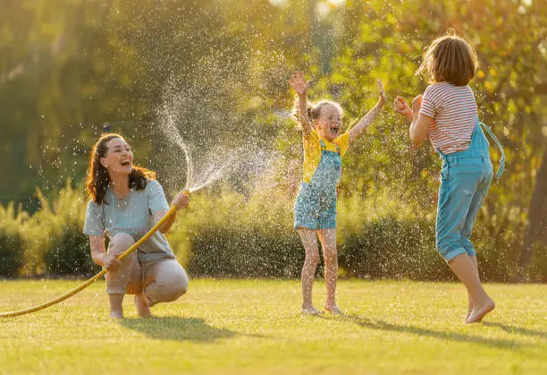 Happy family playing in backyard. Mother sprinkling her kids in hot summer day.