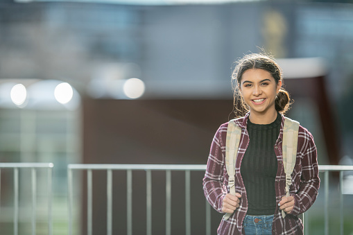 A young female University student of Hispanic decent, poses for a portrait outside the school.  She is dressed in light fall layers and has a backpack slung over her shoulders as she smiles.