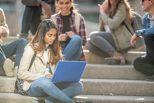 A small multi-ethnic group of University students sit outside on the steps together during break. They are each dressed causally in fall clothing and are chatting amongst each other.  The focus is on the young girl of Indian decent in front, who is sitting cross legged with a laptop out in front of her as she types an assignment up.