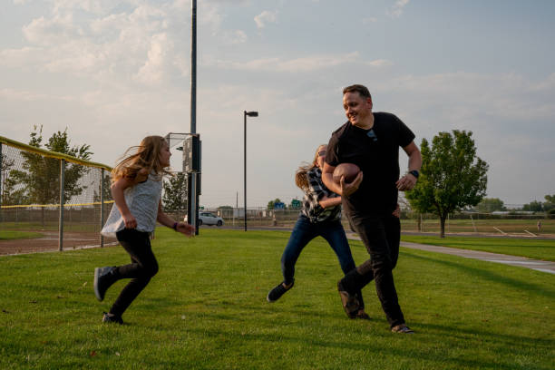 молодой отец играет в футбол со своими двумя дочерьми на травянистом поле в парке поздно вечером летом - american football league стоковые фото и изображения