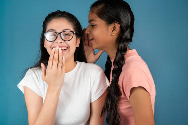 Young woman telling her friend some secret and whispering in her ear. Asian/Indian young woman whispering to her friend ear and telling her some secret or unexpected news. They are gossiping and standing together against isolated blue background. black hair braiding stock pictures, royalty-free photos & images