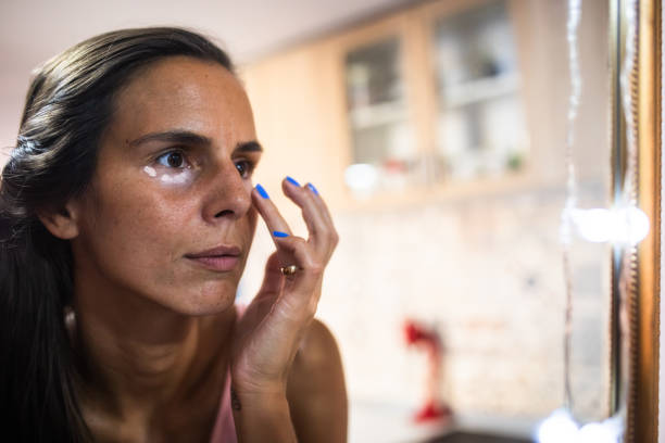 Hide dark circles A woman in her thirties is applying under-eye concealer in front of a mirror using her fingers high collar stock pictures, royalty-free photos & images