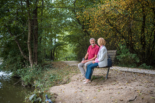 Shot of a senior Caucasian couple enjoying coffee while sitting on a bench next to a lake in a public park. Smiling and  enjoying life.