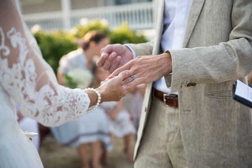 Small intimate wedding ceremony in family beach house. Newlyweds are in their forties. Daughters, witnesses and celebrant are the only guests invited because of COVID. Groom and bride exchanging rings. Horizontal close-up outdoors shot.