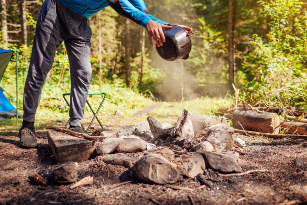 Man extinguishing campfire with water from cauldron in summer forest. Put out campfire. Traveling fire safety rules stock photo