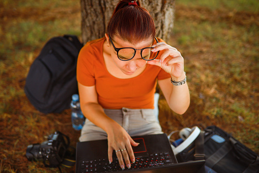Young woman, leaning on the tree in the public park while surfing the net on her laptop