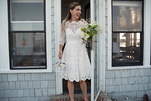 Bride getting out of beach house for small intimate wedding ceremony. Bride is in her forties. There are no other guests invited because of COVID. Horizontal full length outdoors shot with copy space.
