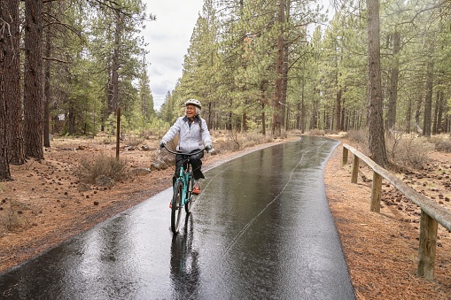 Active mixed race Pacific Islander senior woman smiles while riding her bike along a paved bike path. The environmentally conscious retiree is commuting by bike while vacationing in Oregon. It is a rainy, cool day and the ground is wet from rain. The woman is riding through a wooded rural area.