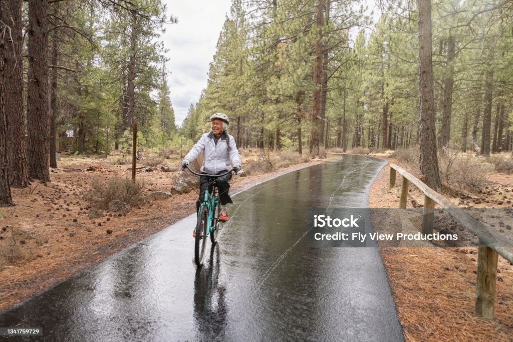 Active mixed race senior woman on relaxing bike ride Active mixed race Pacific Islander senior woman smiles while riding her bike along a paved bike path. The environmentally conscious retiree is commuting by bike while vacationing in Oregon. It is a rainy, cool day and the ground is wet from rain. The woman is riding through a wooded rural area. Cycling Stock Photo
