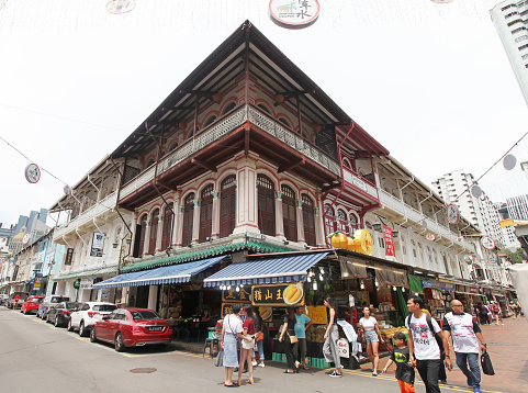 Old colonial shop houses with shuttered windows at the intersection between Temple Street and Trengganu Street in Chinatown. There are parked cars and several people walking in the street and crossing the road.