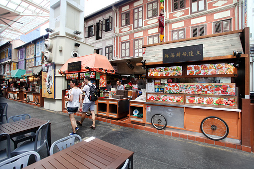 Some of the food stalls in Chinatown Food Street which is a covered street with colonial shop house style buildings with window shutters. Two people are walking past a food stall while a vendor is behind preparing food. Tables and chairs of adjacent restaurants are in the foreground.