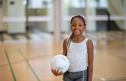 A sweet girl of African decent stands solo in the gymnasium holding a volleyball. She is wearing her athletic gear and smiling .