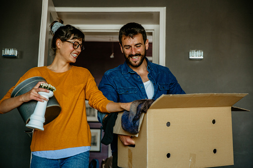 Scandinavian young adult parents, moving into their new place. Entering the living room, with tall gray walls, carrying boxes and stuff in with a big smile and excitement. Loving brunette with eyeglasses and her handsome bearded partner, casually dressed, radiating joy