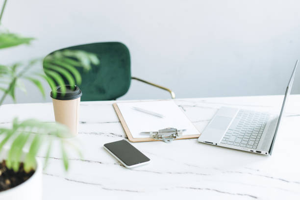 istock Working place with paper cup of coffee, notes, open laptop and smartphone on white marble table in bright modern office room, business and freelance concept 1341790068