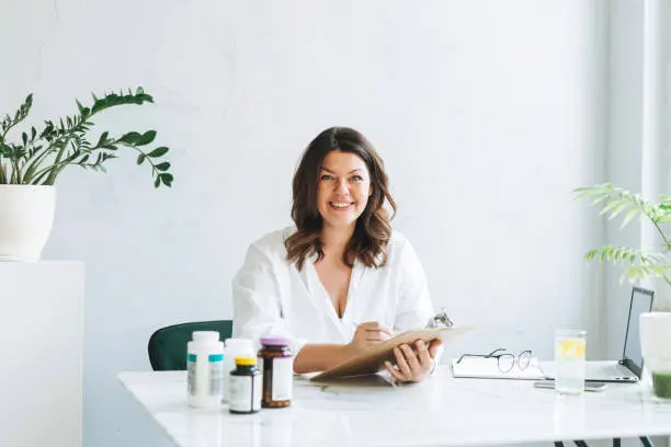 Young smiling brunette woman doctor nutritionist plus size in white shirt working at laptop at modern bright office room. The doctor prescribes prescription for medicines and vitamins at clinic