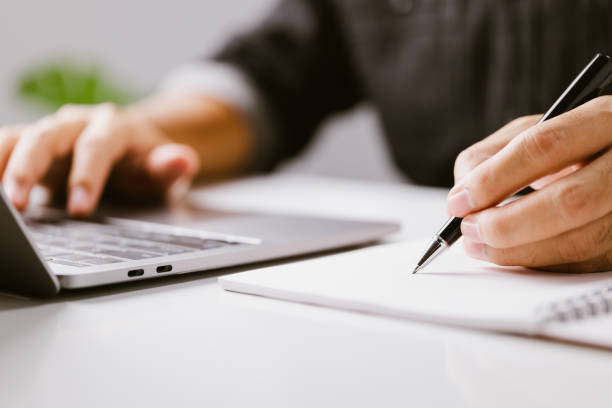 istock Closeup hand of business man writing on note while using laptop computer on desk at home, male planning on note for author and blog, businessman working on table. 1341791715