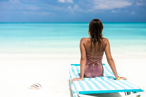 Back view of a woman sitting on a deck chair on the beach and looking at view. Copy space.