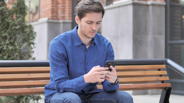 istock Young Man Typing Message on Smartphone, Sitting Outdoor on bench 1341811369