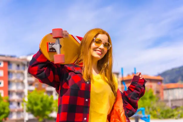 Photo of Skater woman in a yellow t-shirt, red plaid shirt and sunglasses, posing with the board on her shoulder looking at camera