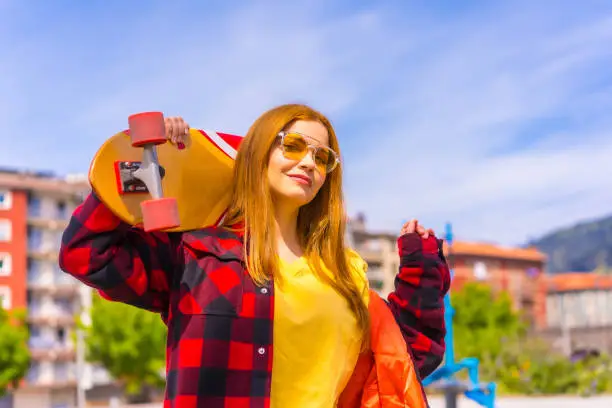 Photo of Skater woman in a yellow t-shirt, red plaid shirt and sunglasses, posing with the board on her shoulder looking at camera