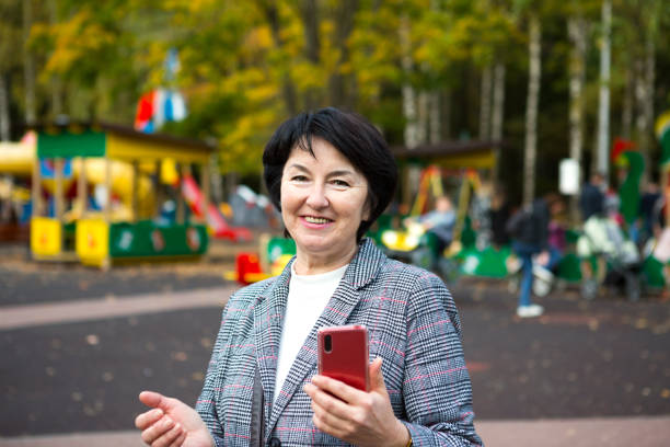 놀이터에 회색 재킷을 입고 가을에 공원에서 50 년 동안 행복한 세노라의 초상화. 한 여성이 웃고 빨간 스마트폰을 사용한다. - women mature adult 50 55 years 50s 뉴스 사진 이미지