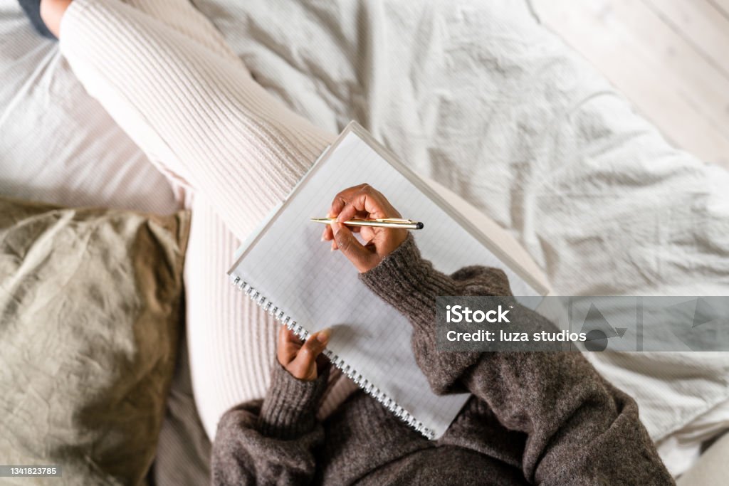 Woman writing in bed A woman is sitting on her bed, using a note pad to write. She is listening to something on her headphones. Diary Stock Photo