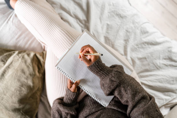 Woman writing in bed A woman is sitting on her bed, using a note pad to write. She is listening to something on her headphones. diary stock pictures, royalty-free photos & images